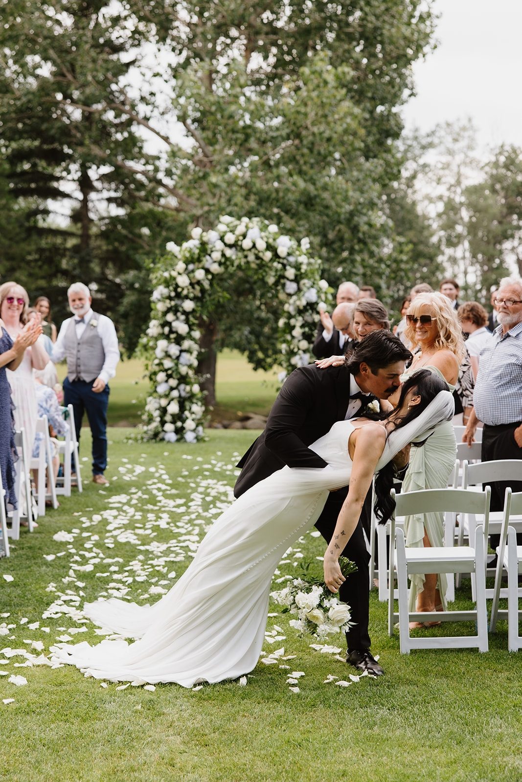 Luxury Wedding ceremony at Hilltop wedding center featuring a full round top archway with smilax and greenery accented by white flowers, white roses as well as a bride being dipped by the groom holding a bride bouquet and white rose petals on the grass