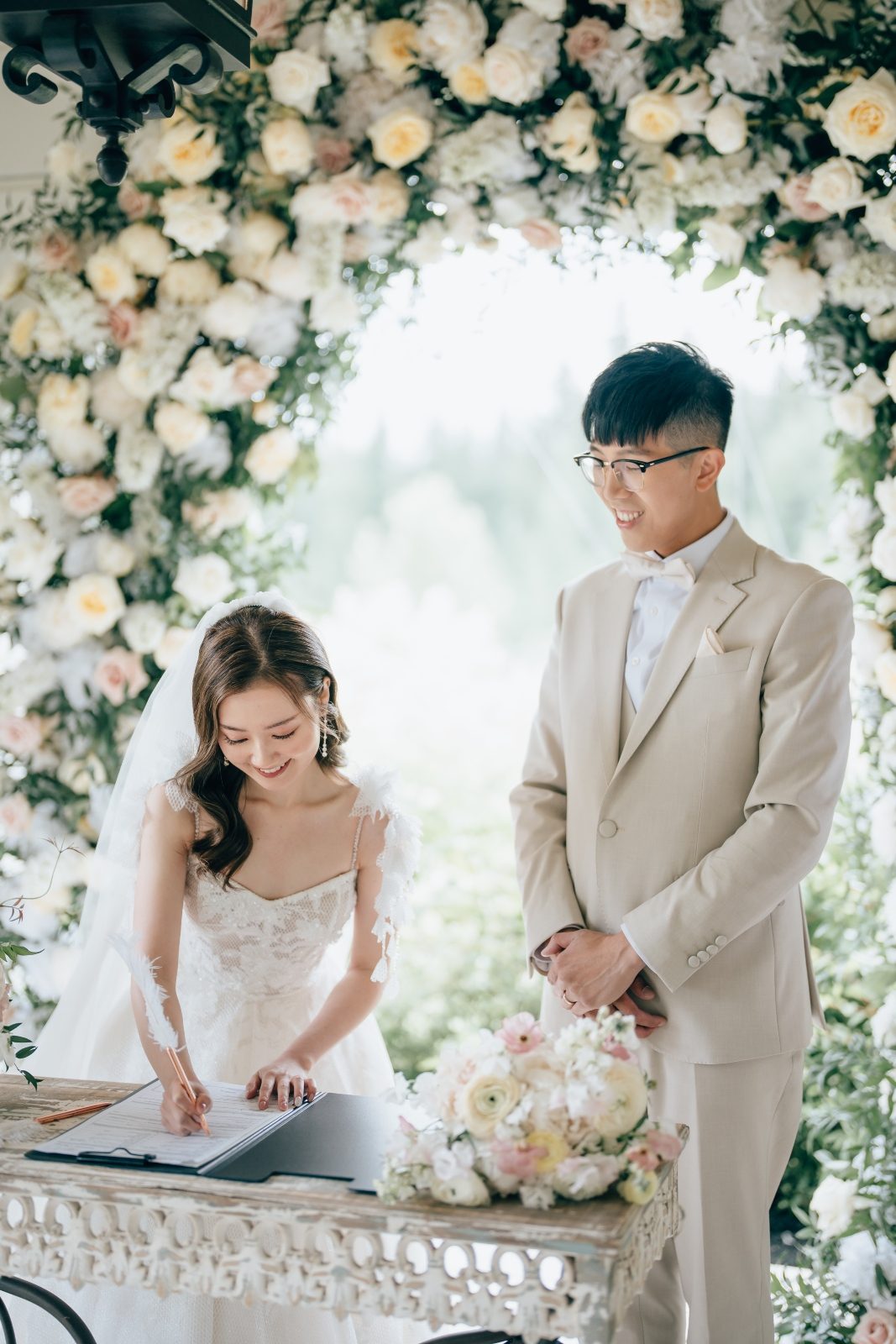 Gorgeous wedding bride and groom in a wedding dress at the signing table with her bouquet designed with delicate pastel blooms in white and blush including ranunculus, butterfly ranunculus and garden roses and a luxury wedding arch behind them at Archway Manor