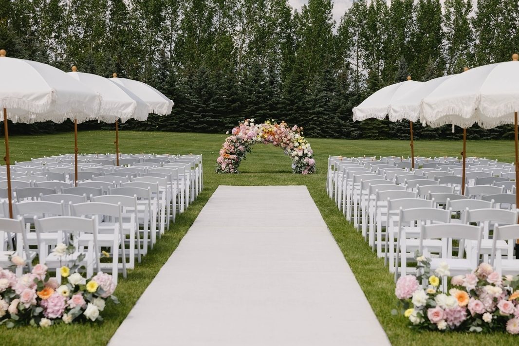 Wedding ceremony low arch designed in an ombre style of spring bright pastels in yellow, peach coral pink and white with lush florals including gerbera daisy and hydrangea with umbrellas and floral aisle hedge aisle markers at Tangled Roots venue
