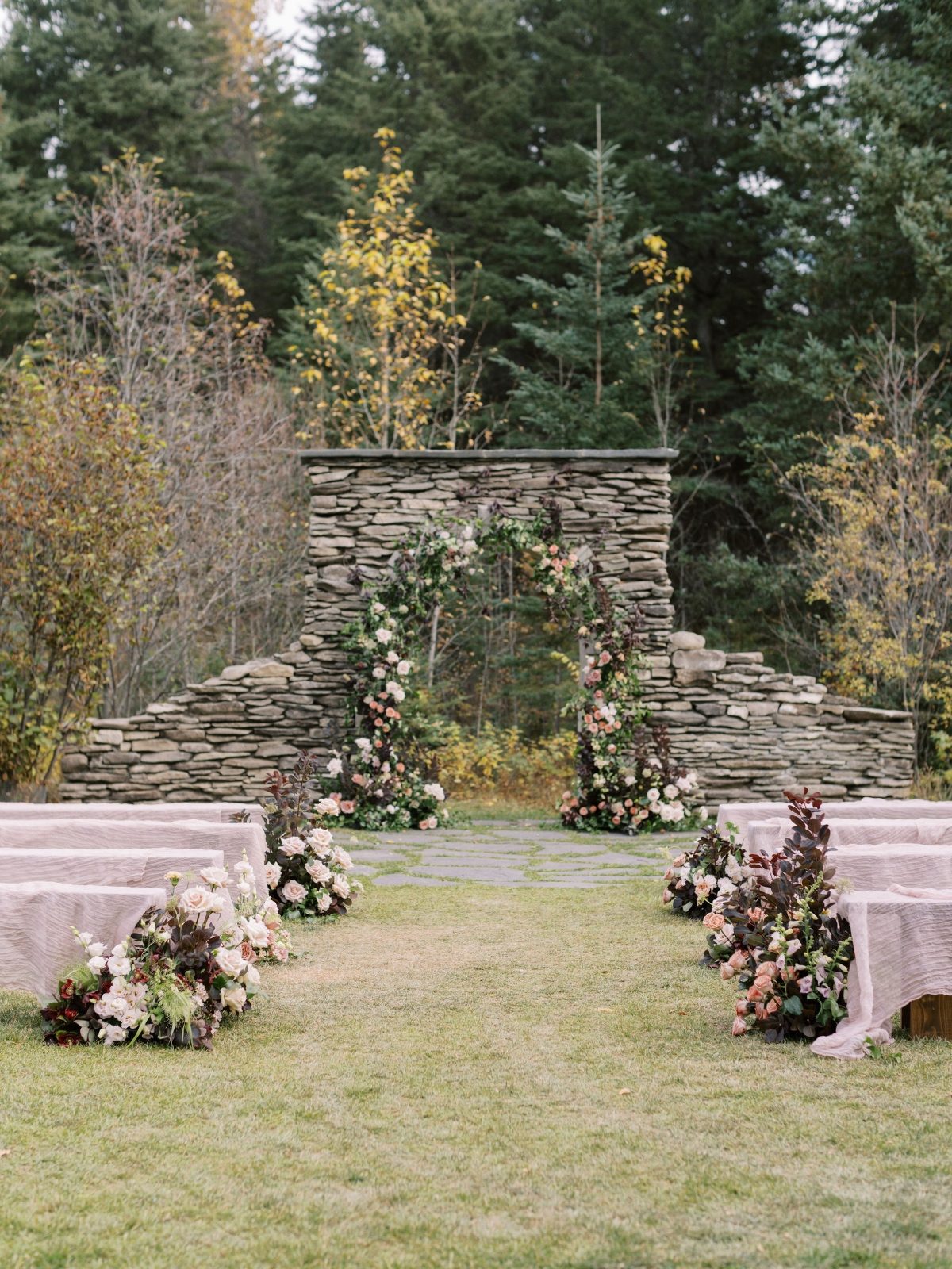 Wedding ceremony set up with wild and natural stone arch and aisle hedges alsi markers, flowers in burgundy, blush, terra cotta, white and greenery at The Valley Weddings. Ceremony benches with pink cheesecloth.
