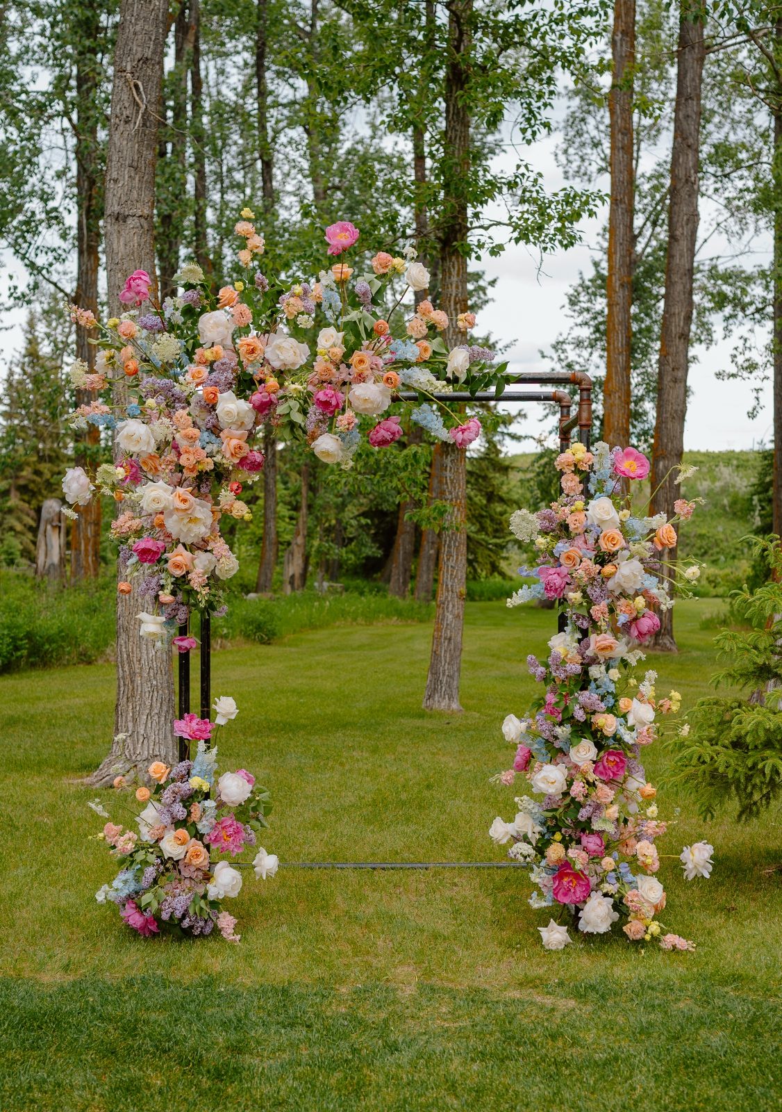 An outdoor spring wedding pipe archway designed with bright pastel flowers including peonies, ranunculus, delphinium, tweedia, delphinium and lilacs and garden roses at 52 North Venue