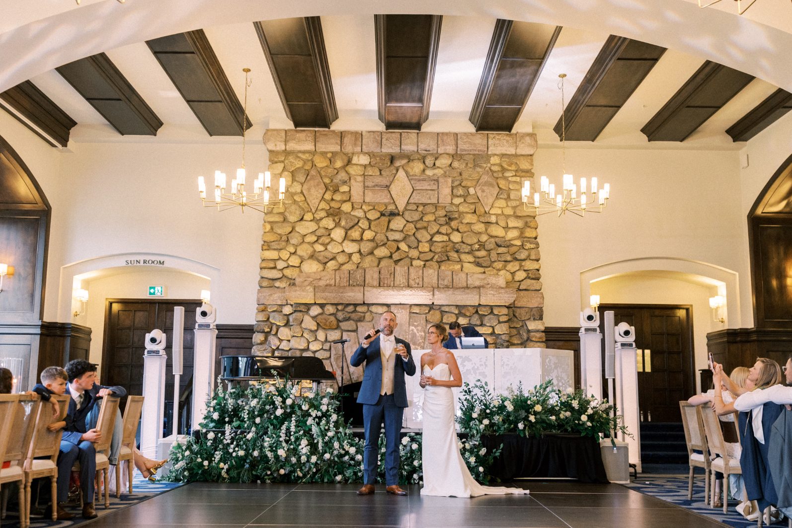 Reception Bride and groom giving speeches in front of dramatic luxury wedding floral installation and grand stone fireplace.