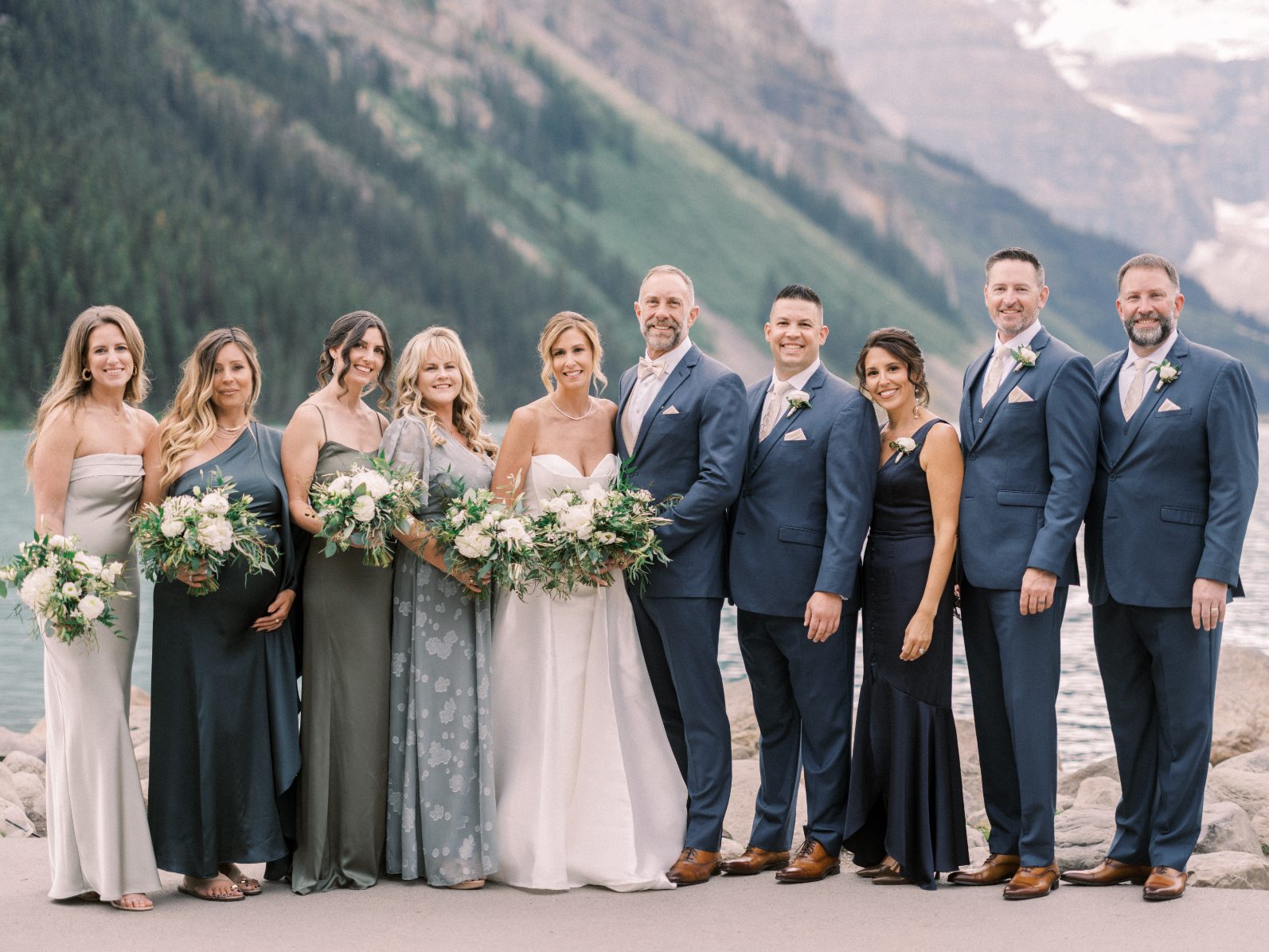 Wedding Party Full wedding party gathered along a serene mountain lake, featuring coordinated navy and sage attire with elegant white bouquets.