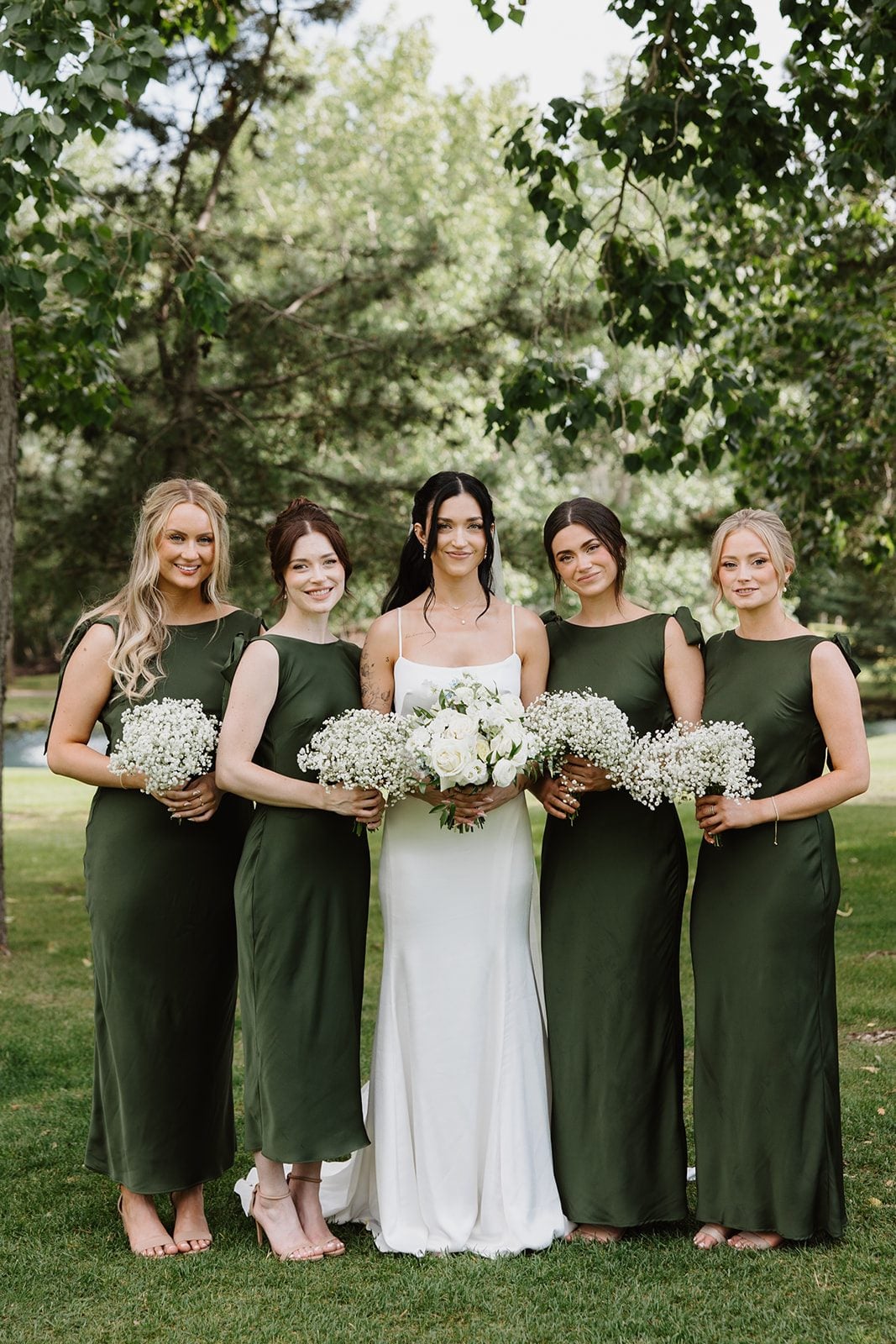 Cassidy + Adon The bride holds a white bridal bouquet and stands with her bridesmaids in green satin dresses holding bouquets of baby's breath.