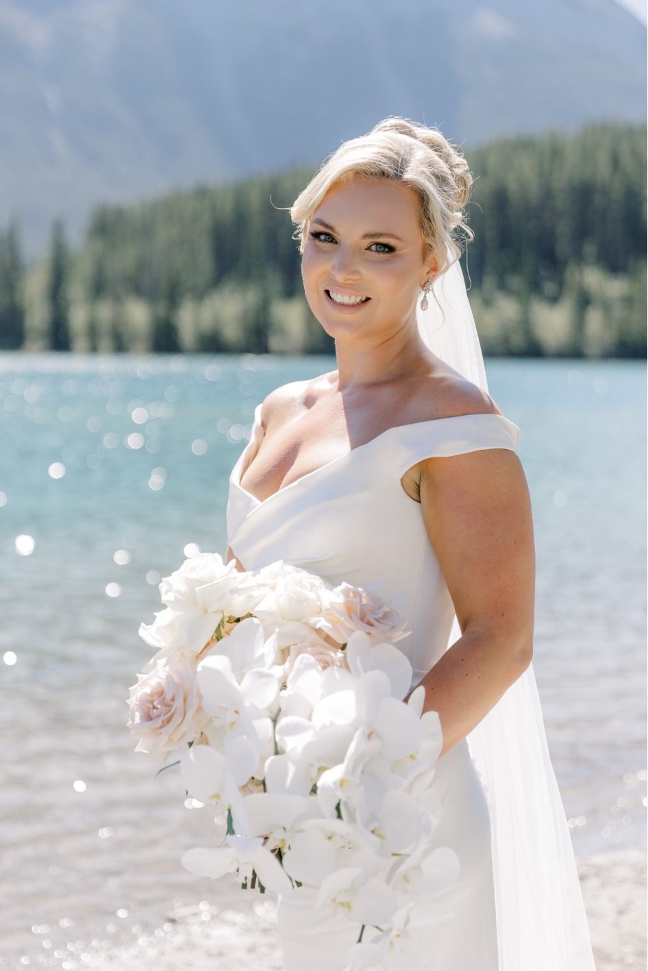 Blushing Bride Bride stands in front of sparkling blue waters of a mountain lake, smiling at the camera holding her bouquet.
