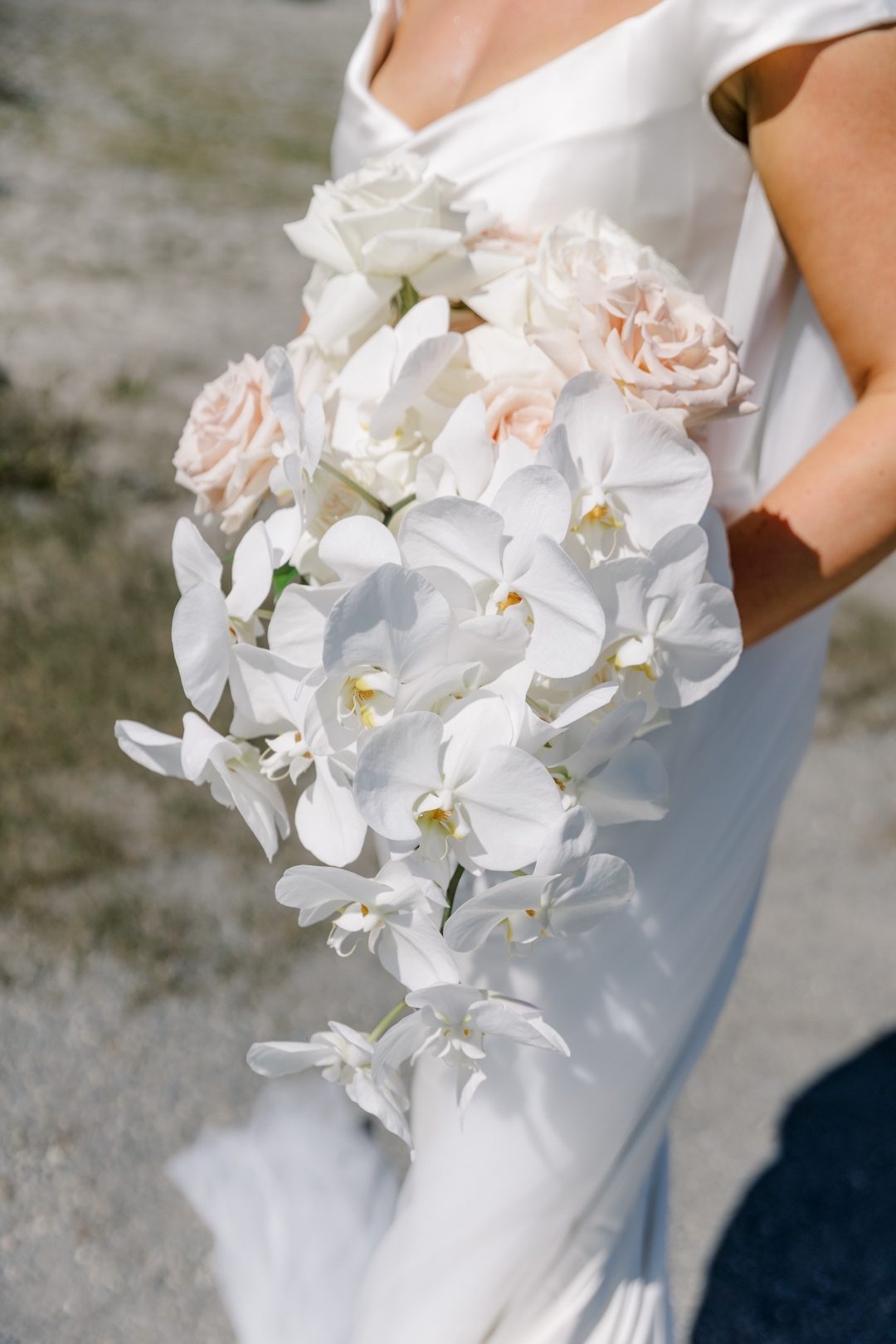 Bouquet Bride holds a cascading bouquet of white orchids and soft blush roses against a natural outdoor backdrop.
