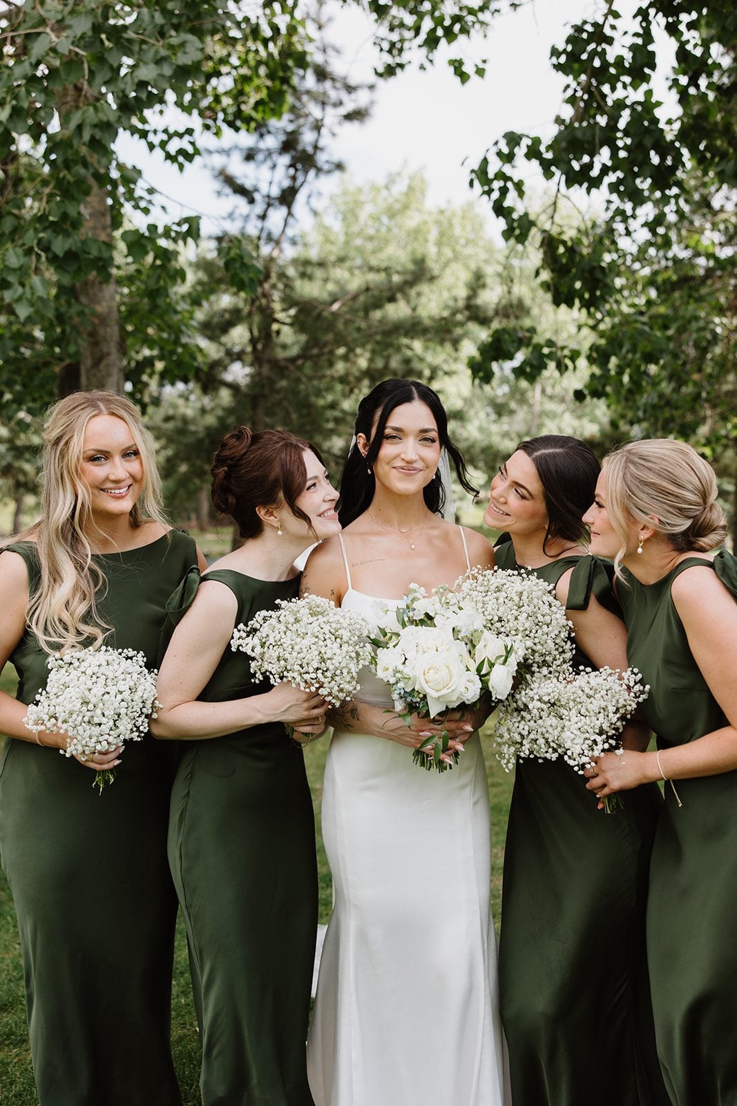 Cassidy + Adon Bride stands smiling holding a white bridal bouquet while her bridesmaids, wearing green satin gowns and holding baby's breath bouquets, look at her smiling.