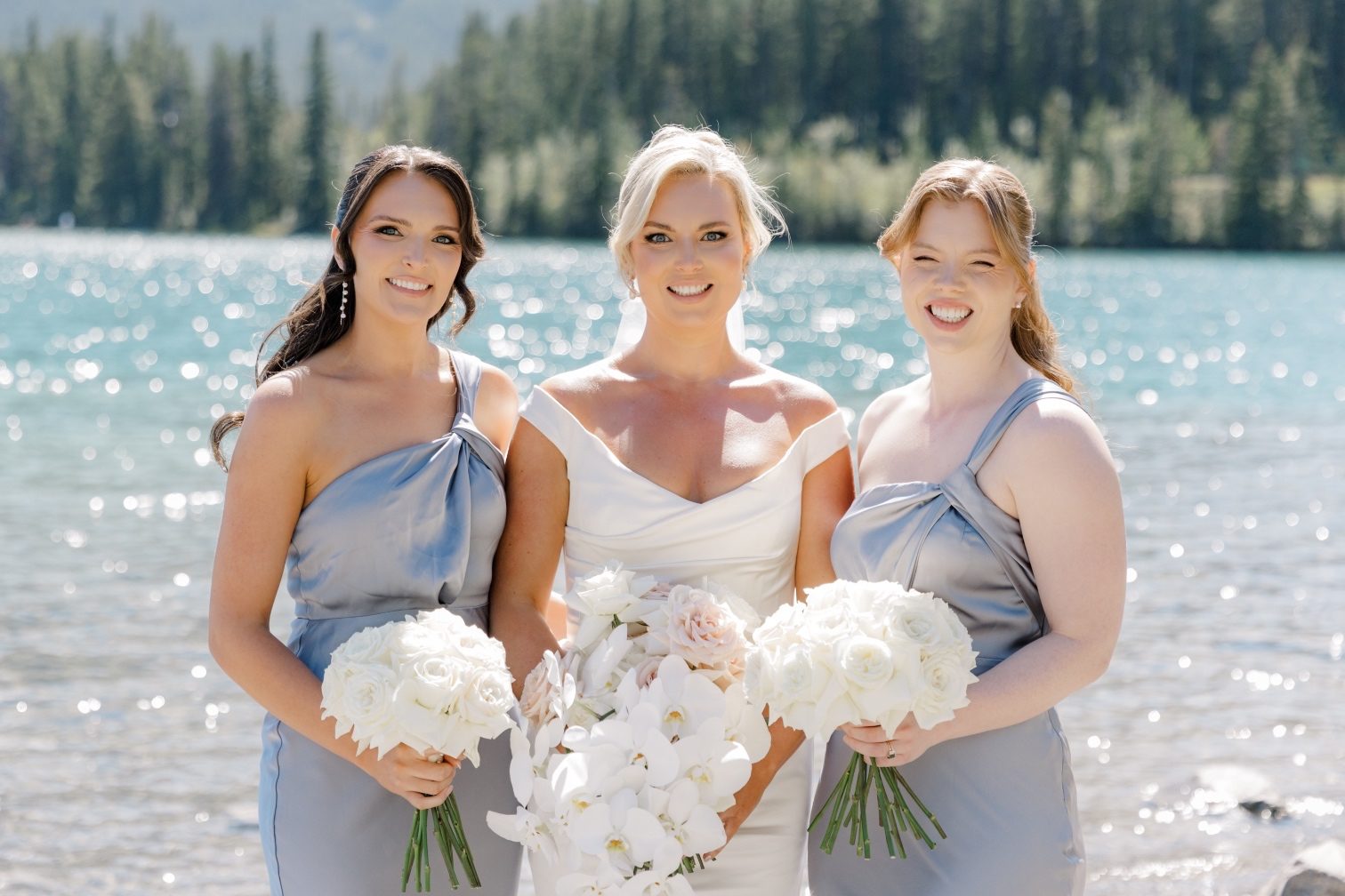 Bride + Bridesmaids Close up of bride and bridesmaids holding white rose bouquets while standing at the edge of Lake Louise.