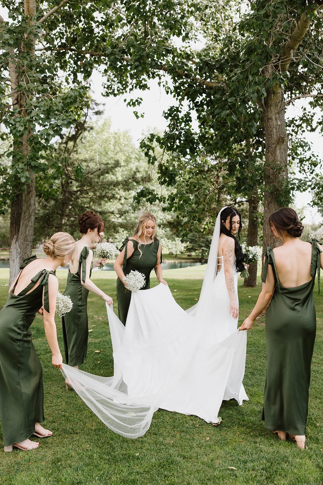 Cassidy + Adon Bridesmaids in green satin dresses, holding baby's breath bouquets help adjust the brides dress and veil.