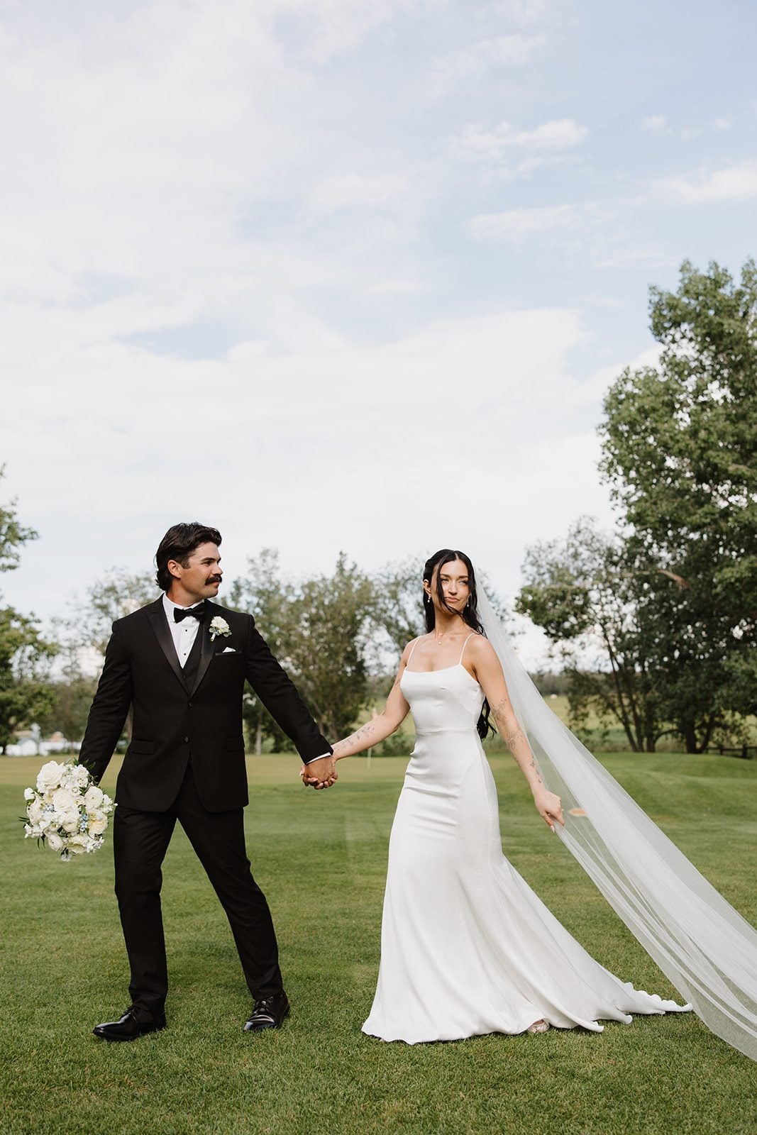 Cassidy + Adon Bride and groom walk hand in hand across Hilltop Wedding Center's beautiful grounds, holding a white bridal bouquet.
