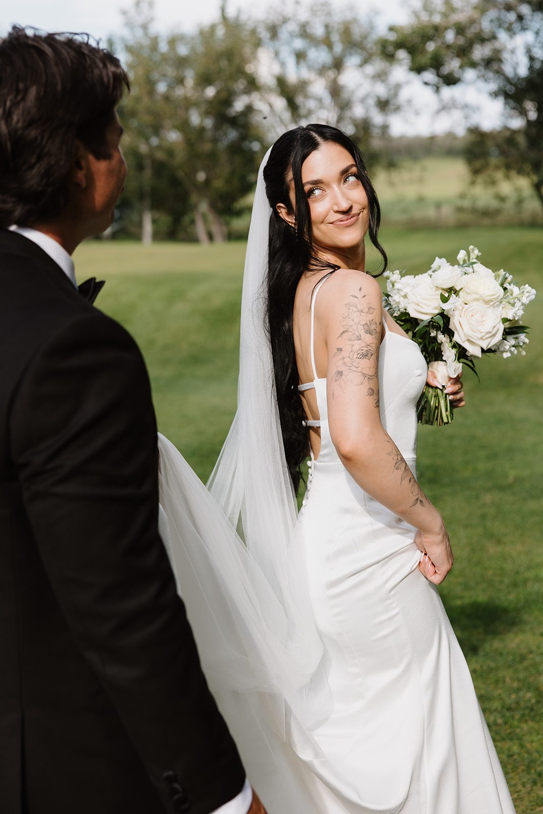 Cassidy + Adon Bride holds a white bridal bouquet of roses, ranunculus, sweet peas, tweedia, and eucalyptus, looking over her shoulder at the groom who is holding her veil.