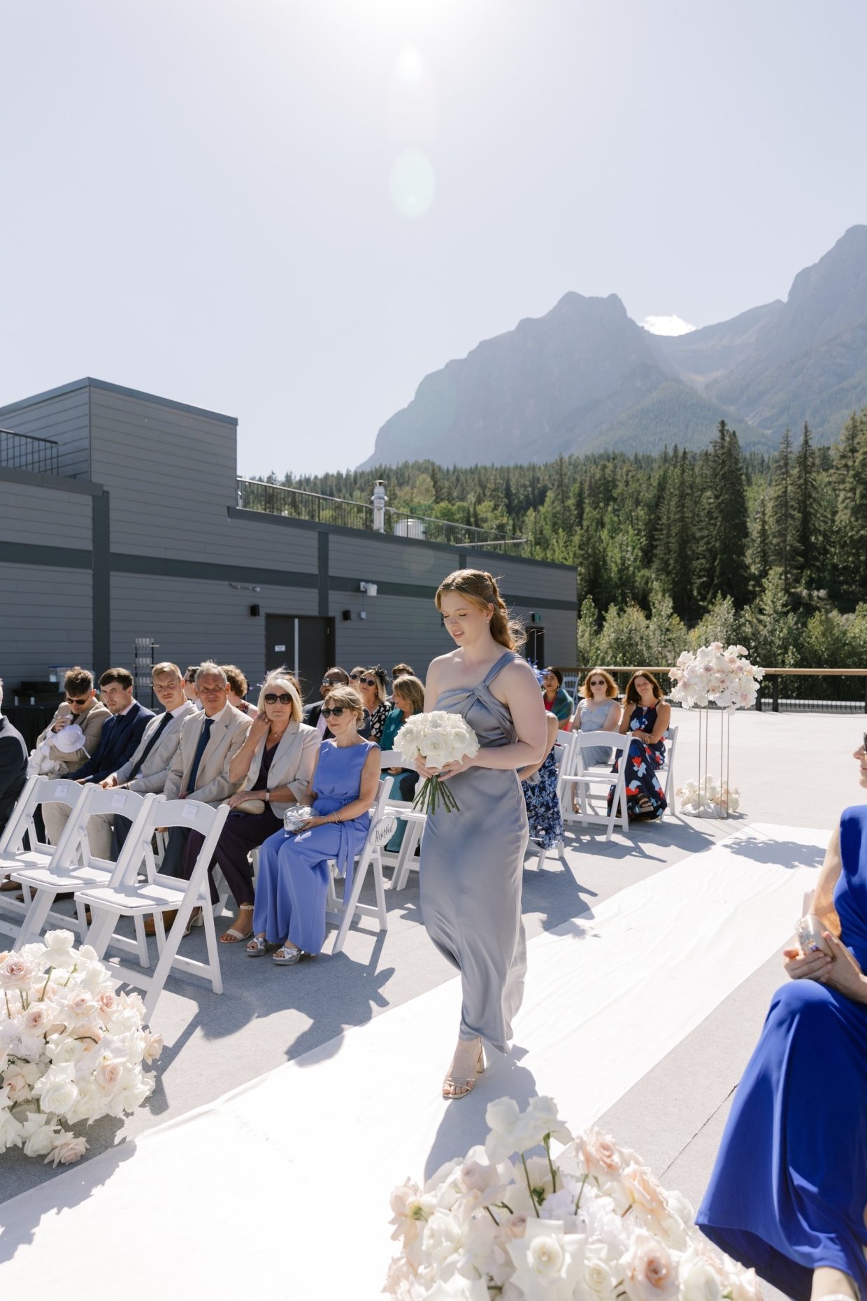 Bridesmaid Bridesmaid walking down the aisle holding a white rose bouquet with guests seated behind her and mountain views surrounding the ceremony.