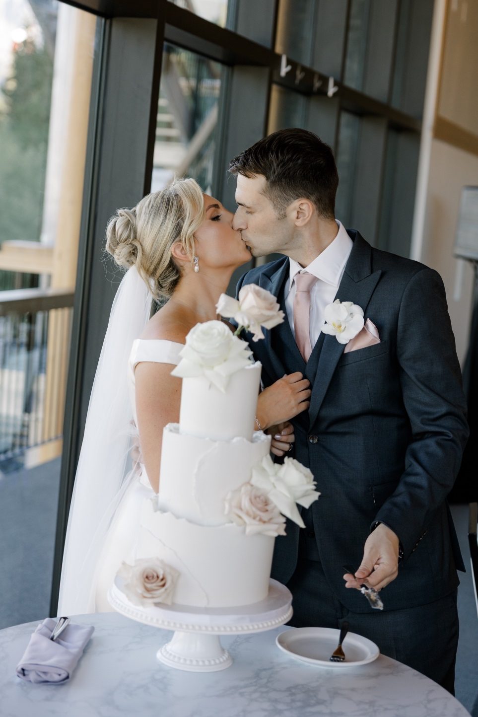 Cake Cutting Bride and groom share a kiss while cutting their white three tier wedding cake adorned with blush and ivory roses at their mountain view reception.