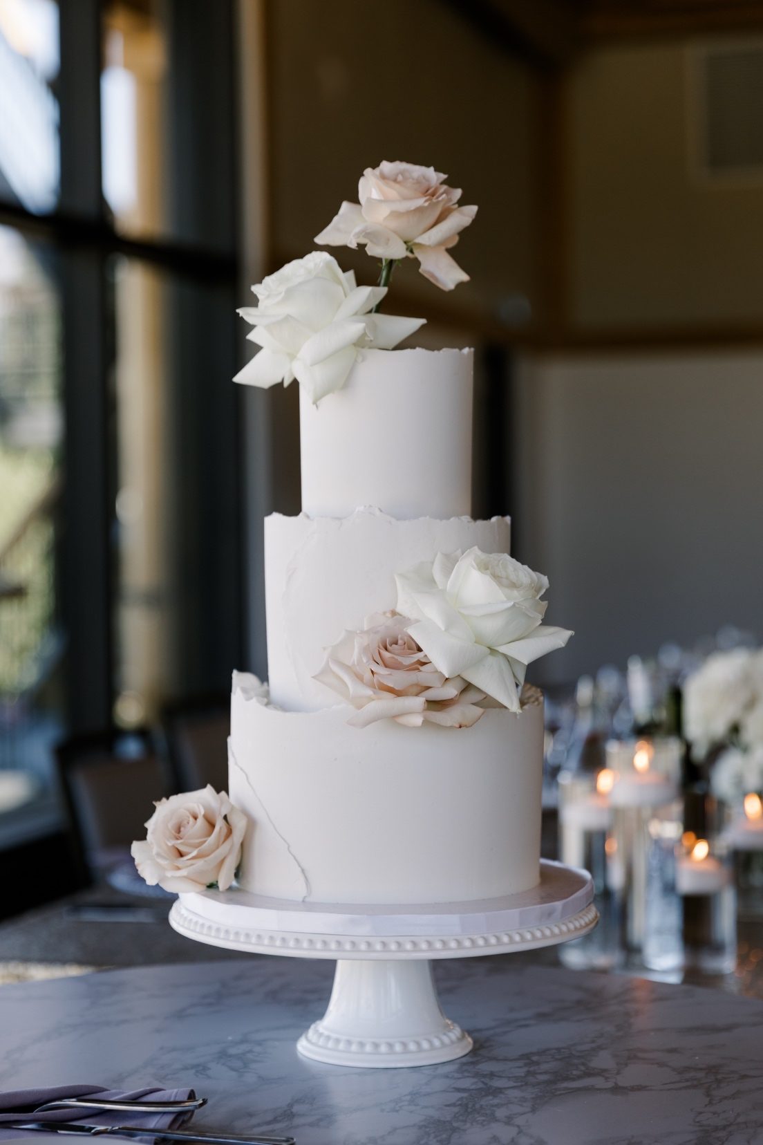 Cake Flowers Three tiered wedding cake with soft blush and ivory roses, displayed on a white pedestal cake stand inside a modern mountain view reception space.