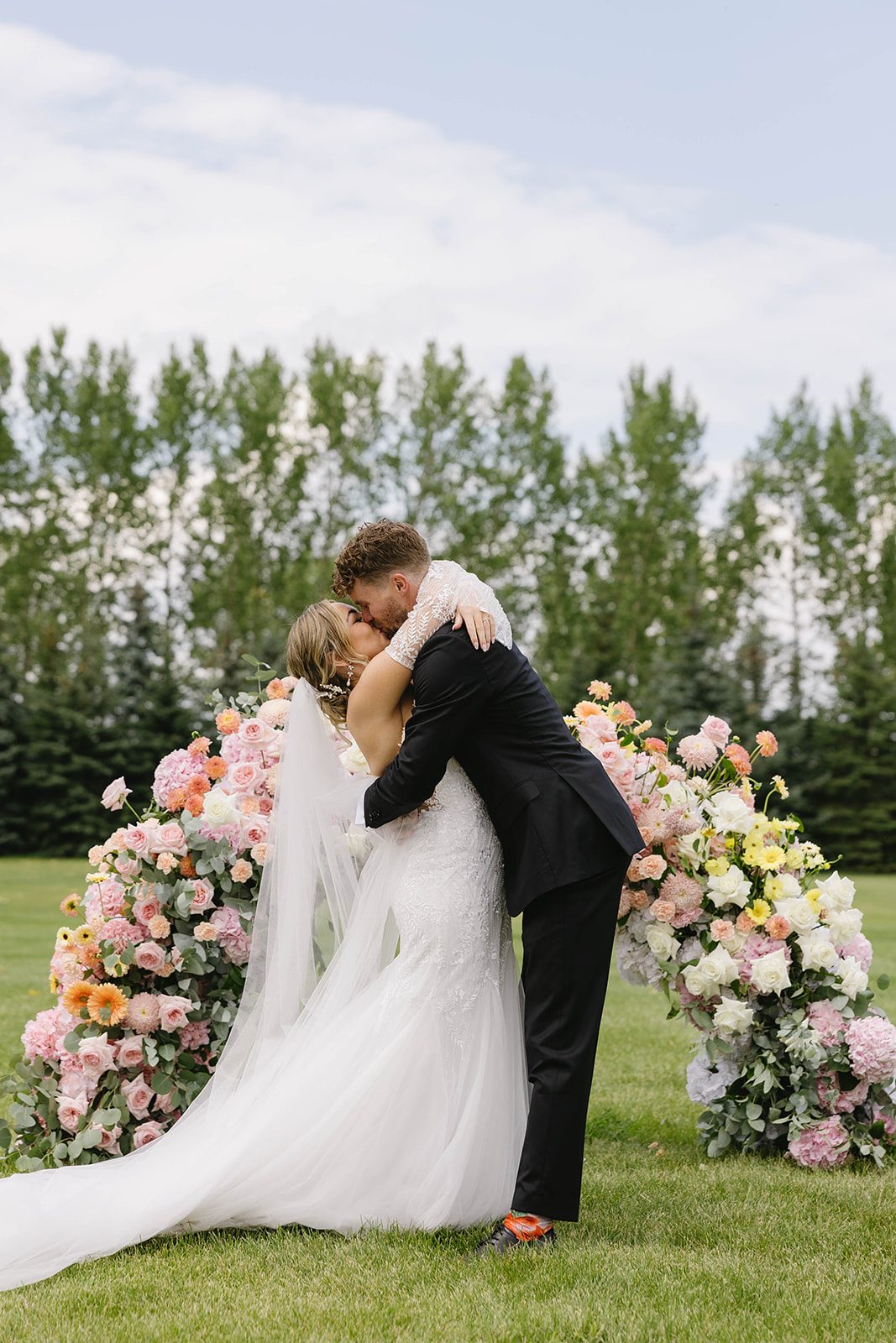 Bride and groom share a kiss after saying "I do" in front of a low, ombre, pastel floral arch at their Tangled Roots ceremony.