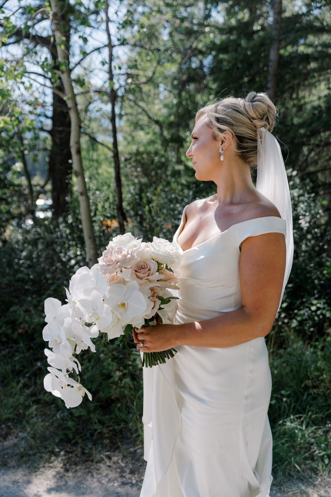 Cascade Bride holds a cascading bouquet while standing among trees near the lake, sunlight on her dress.