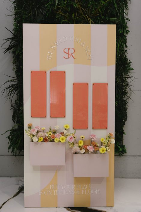 A colourful seating chart with a blush pink and peach backdrop, orange list of tables, and two floral boxes with pastel arrangements of dahlias, gerbera daisies, lisianthus, ranunculus, roses, carnations, and stock.