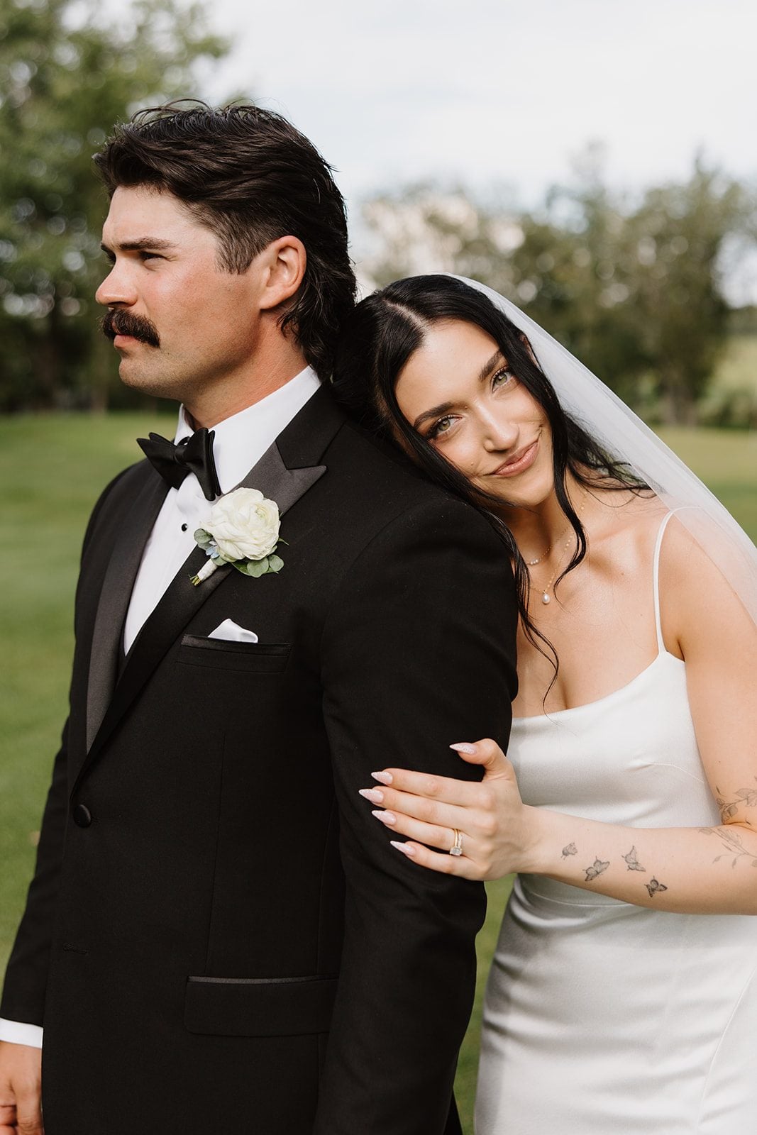 Cassidy + Adon Bride and groom take portraits at Hilltop, groom with a white boutonnière looks into the distance while bride leans on his shoulder looking at the camera with a soft smile.