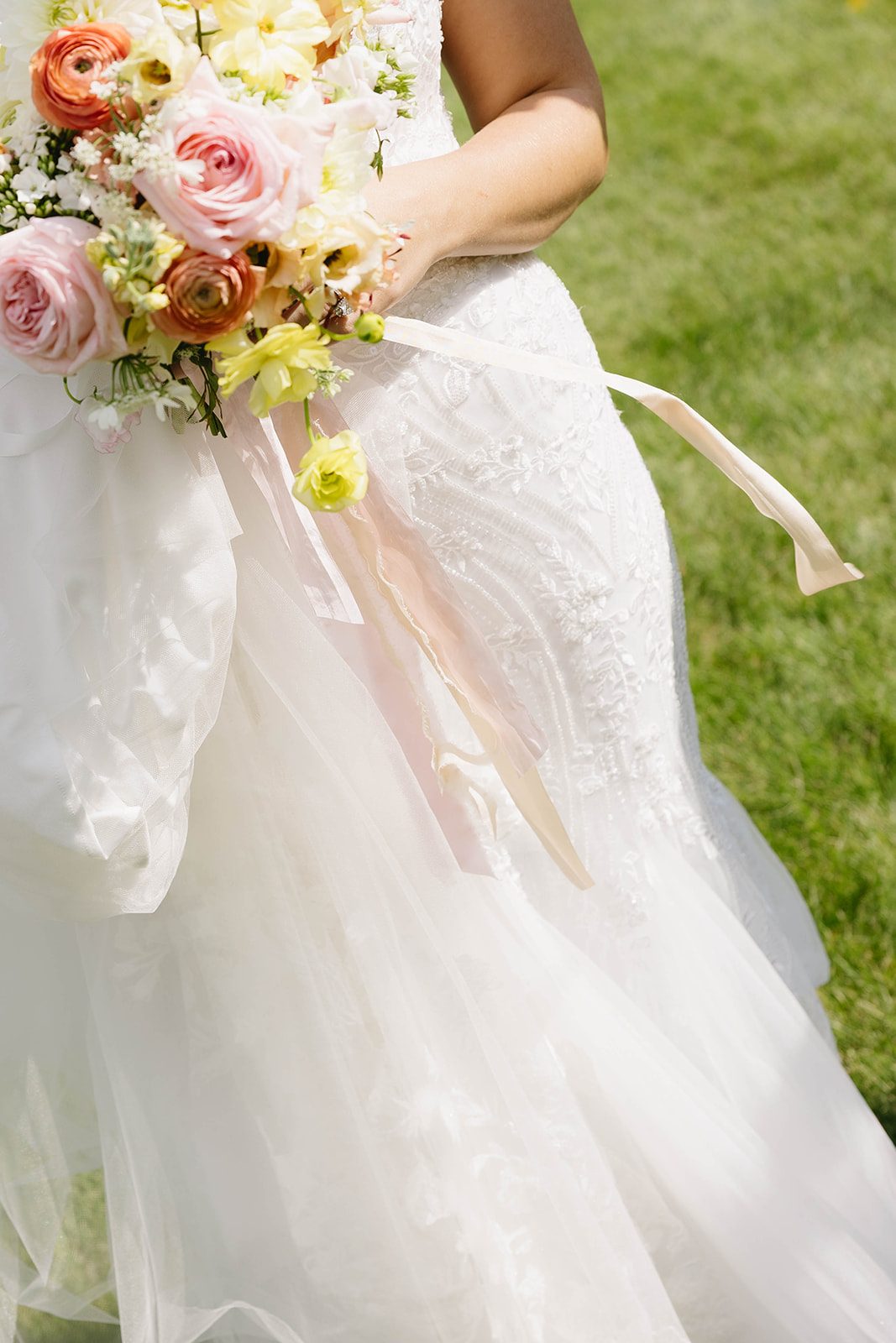 A bride holds a bouquet of garden roses, dahlias, lisianthus, stock, ranunculus, sweet peas and orlaya in blush pink, peach, butter yellow with trailing silk ribbons.