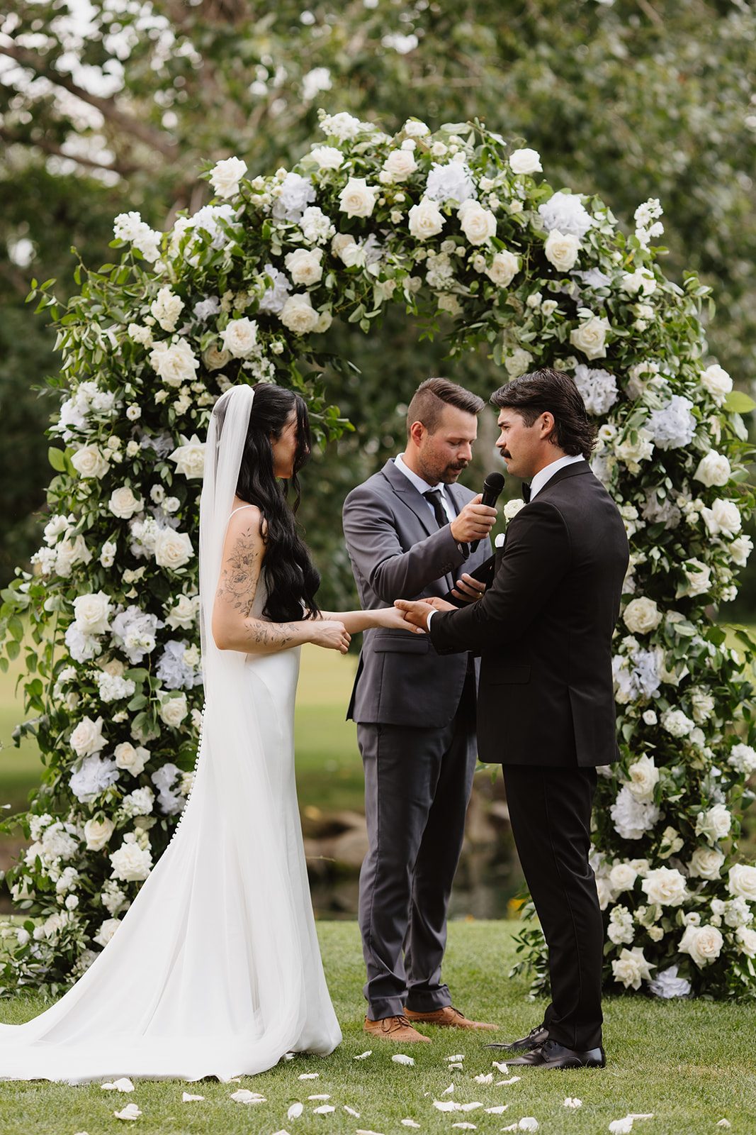 Cassidy + Adon The bride and groom stand in front of a lush green and white floral arch during their Hilltop ceremony, saying their vows.