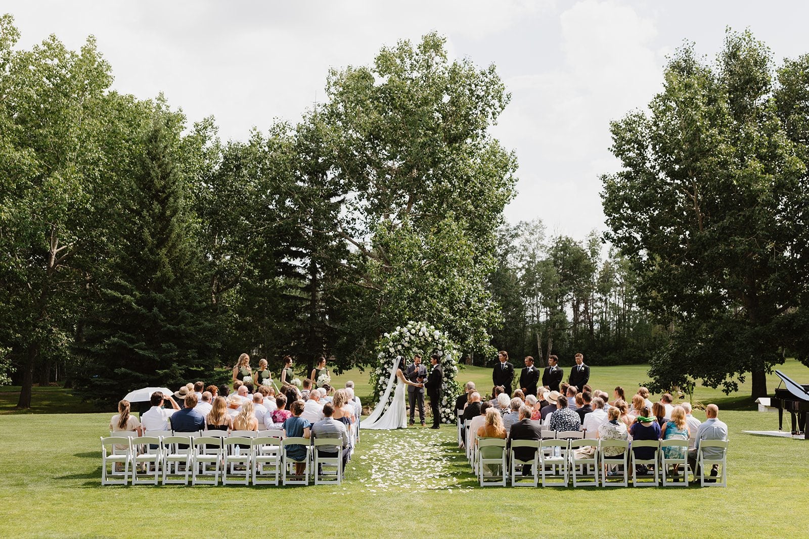 Cassidy + Adon A zoomed out shot of this green and white wedding ceremony at Hilltop wedding centre with guests sitting in rows of white chairs while the bride and groom sat their vows in front of a traditional, lush, green and white floral arch.