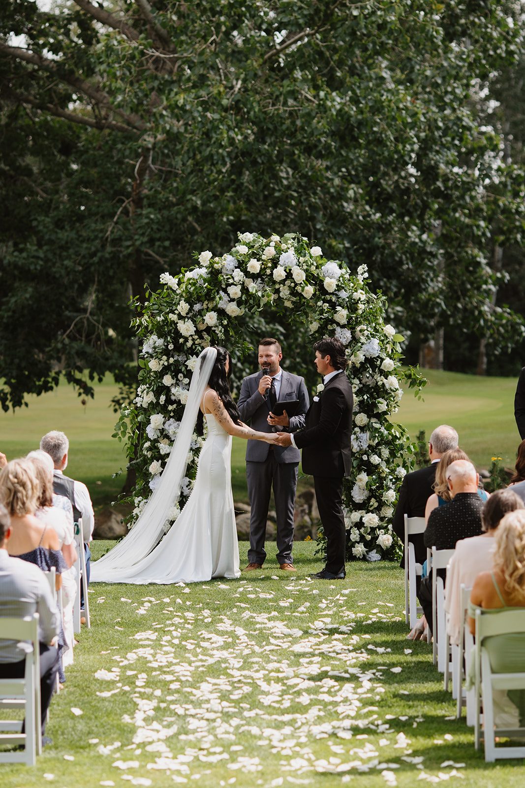 Cassidy + Adon Bride and groom stand in front of their lush green and white floral arch during their ceremony at Hilltop, hands clasped gazing at each other lovingly.