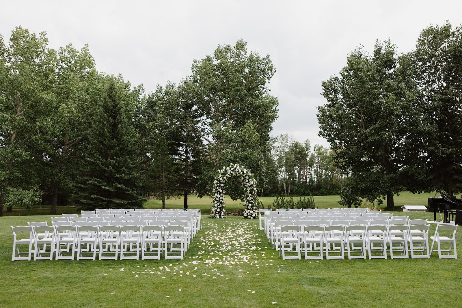 Cassidy + Adon The ceremony set up at Hilltop Wedding Center for this green and white wedding, with tidy rows of white chairs, white rose petals scattered down the aisle and a large, traditional, solid arch full of white flowers and lush greenery for a very romantic look.