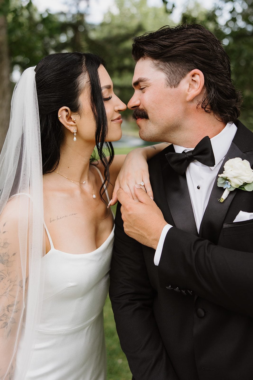 Cassidy + Adon Bride and groom (with a white boutonniere) smiling at each other nose to nose in front of the pond at Hilltop.
