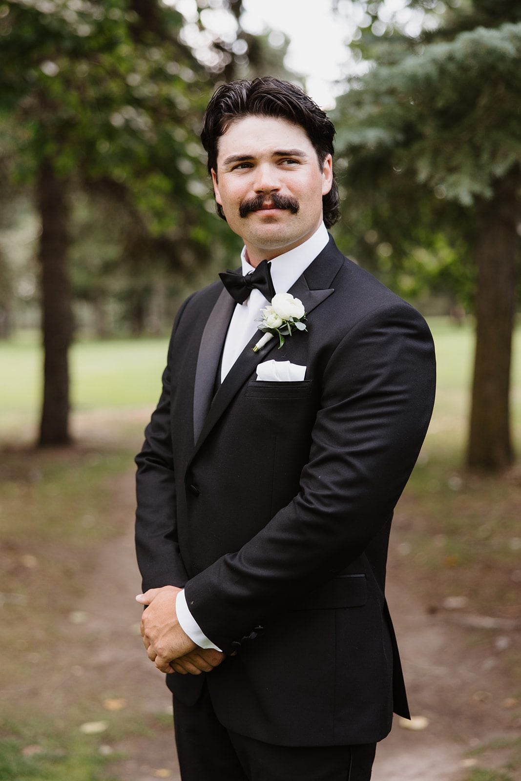 Cassidy + Adon Groom wearing a black tuxedo wears a white boutonnière with ranunculus, spray rose and tweedia accent, smiles softly with his hands together.