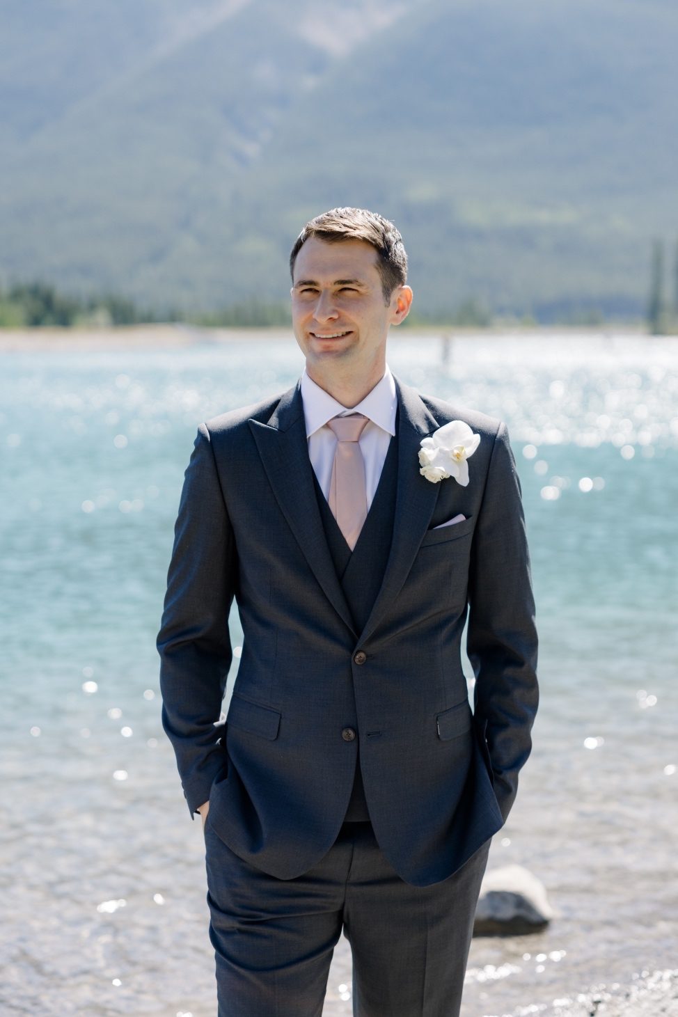 Groom Groom smiling at the lakeshore in a charcoal suit with a blush tie and white orchid bloom boutonnière.