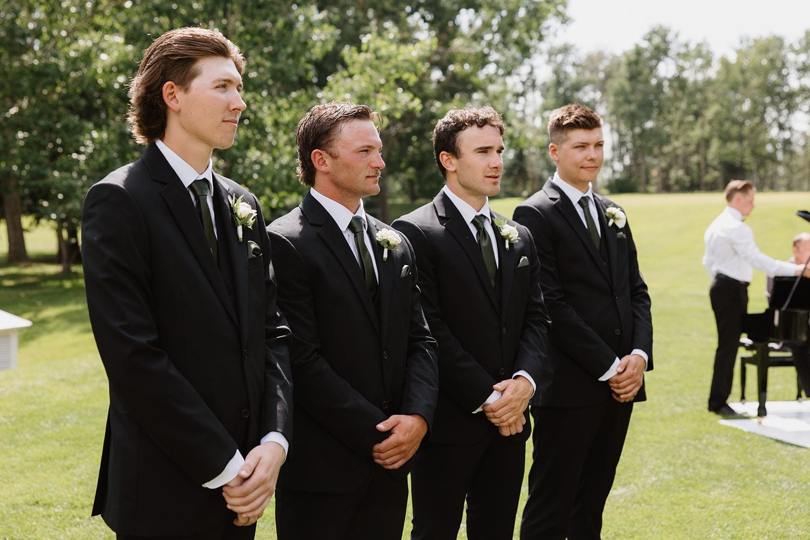 Cassidy + Adon Groomsmen stand in a row in their black suits during this green and white wedding ceremony at Hilltop, wearing white spray rose and ranunculus boutonnières.