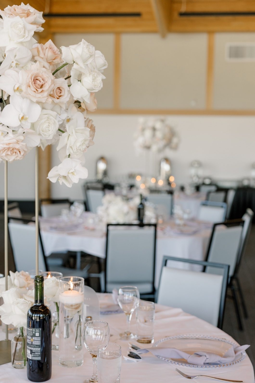 Guest Tables Tall flora centrepiece of ivory and blush roses with cascading orchids on a round guest table set with elegant glassware and soft grey napkins.