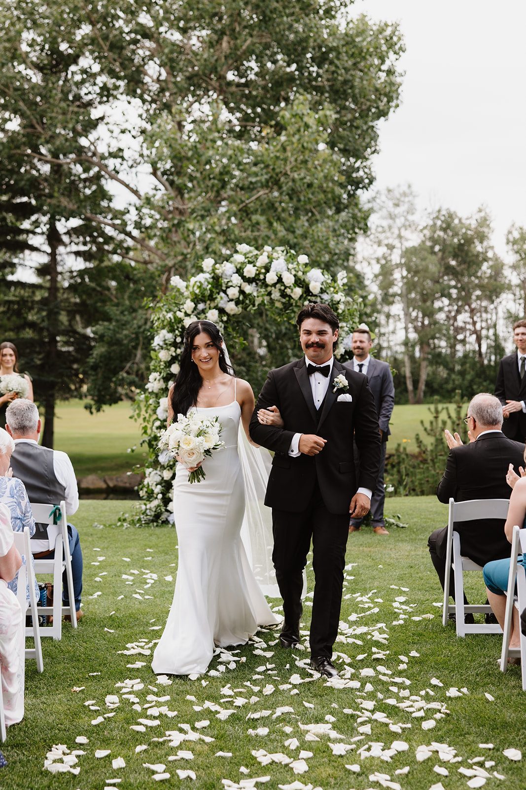 Cassidy + Adon Bride and groom exit the ceremony down an aisle of scattered white rose petals with a lush green and white floral arch behind them.