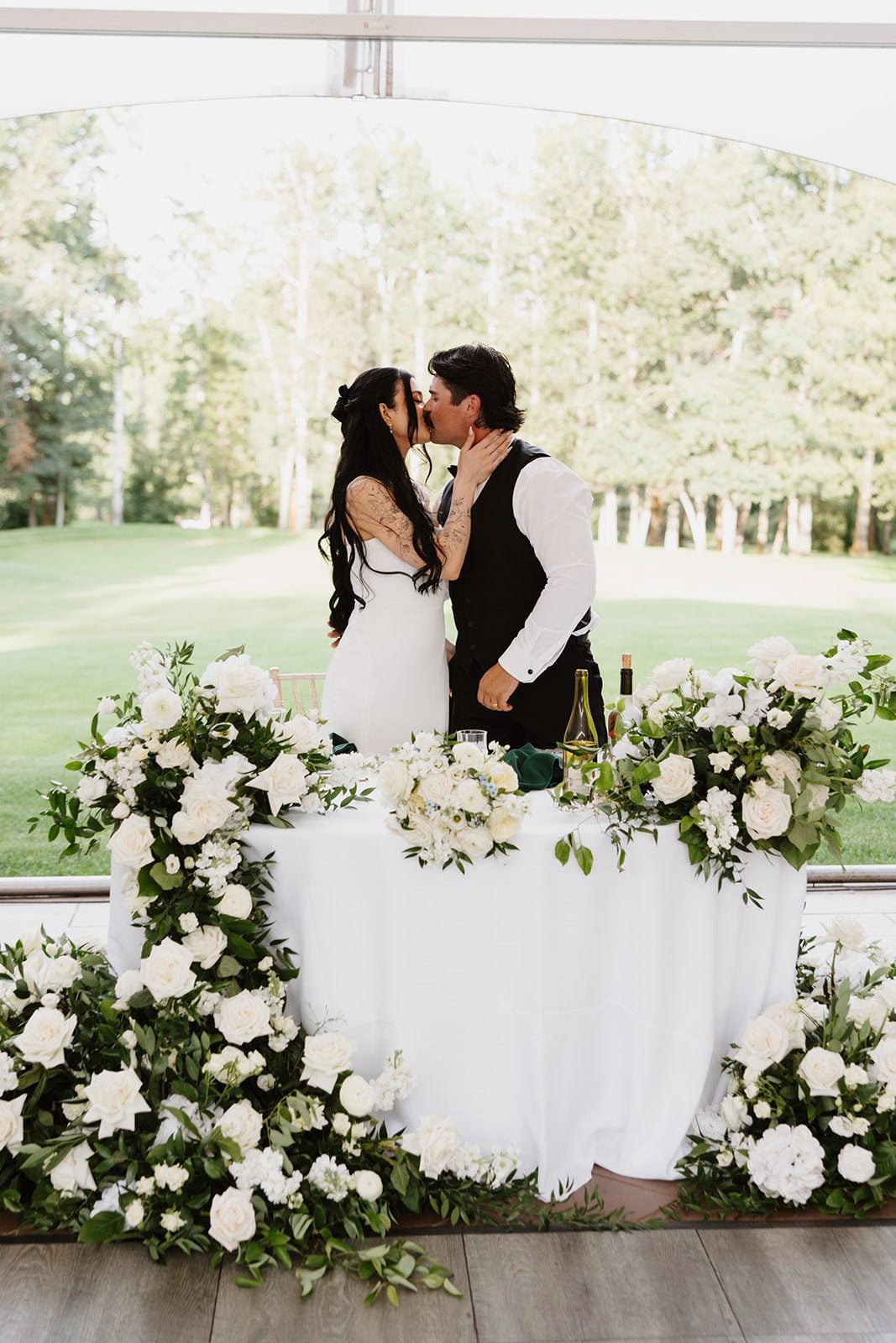 Cassidy + Adon Bride and groom share kiss behind their sweetheart table at Hilltop, beautifully decorated with lush green and white arrangements.