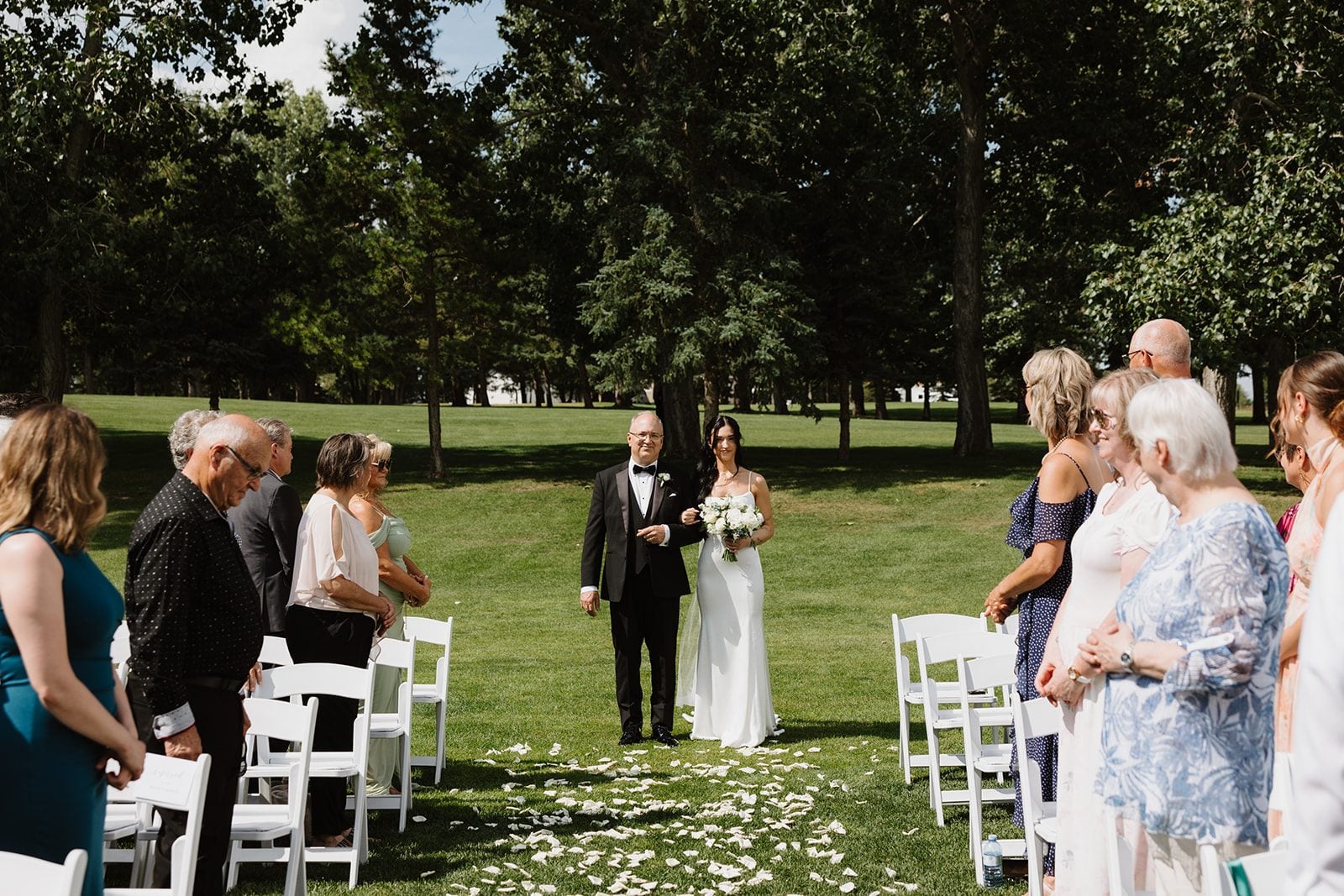 Cassidy + Adon The bride starting down the aisle off white rose petals, on her father's arm while holding a beautiful white bridal bouquet of roses, ranunculus, sweet peas, accents of tweedia and eucalyptus.