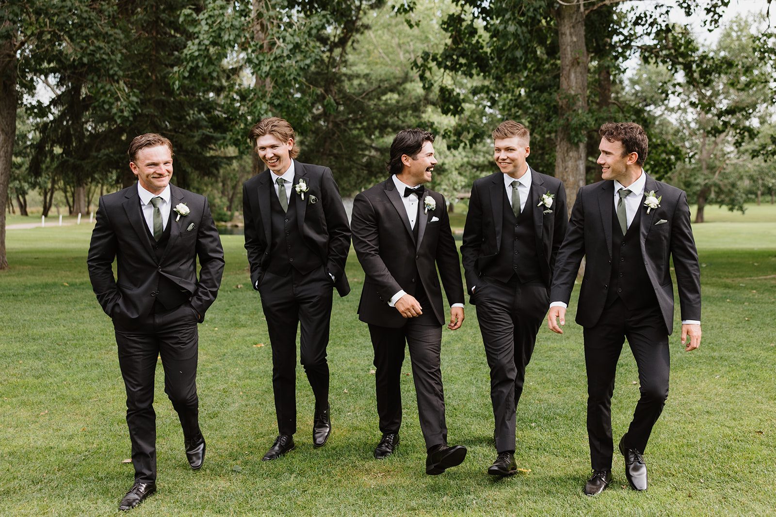 Cassidy + Adon Groom and groomsmen walking together on the grounds at Hilltop wearing black suits and white boutonnières of ranunculus and spray roses.
