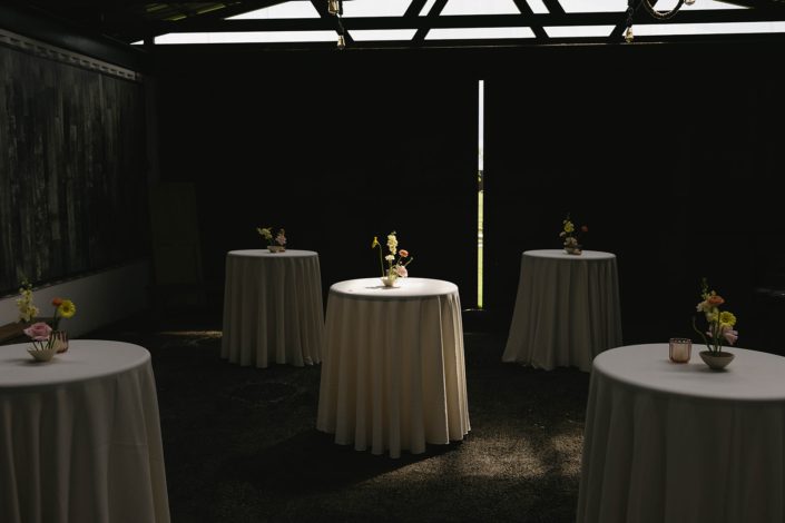 A grouping of cocktail tables with ivory linens and an ikebana styled arrangement featuring a gerbera daisy, ranunculus, rose and stock, with the table in the centre in a sun beam.
