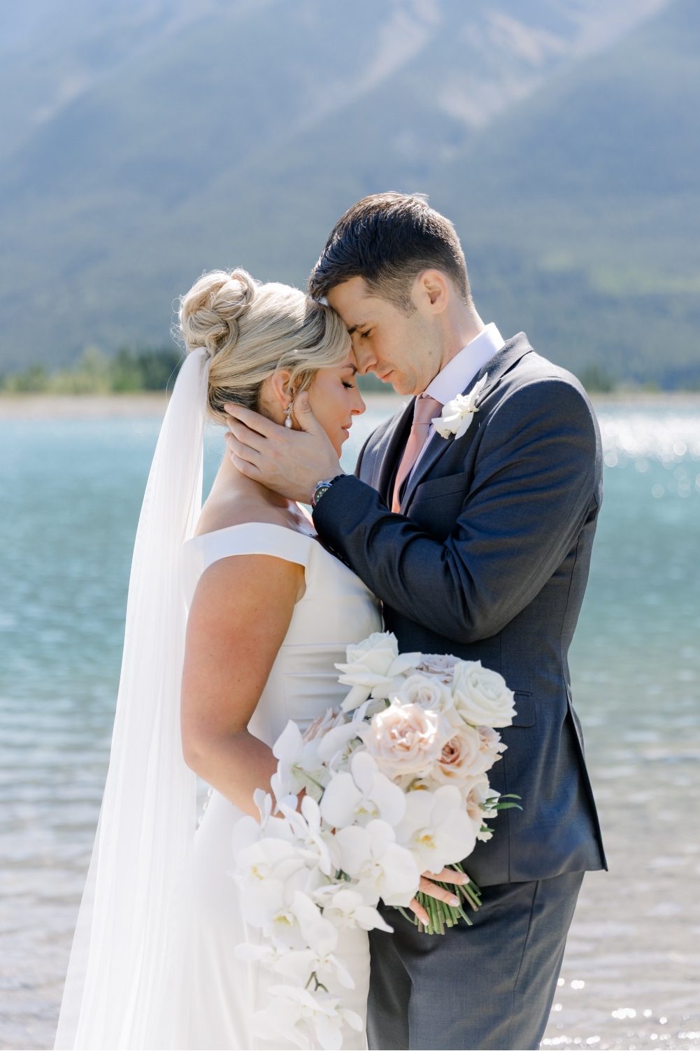 Love Bride and groom embracing gently at the water's edge with mountains behind them.