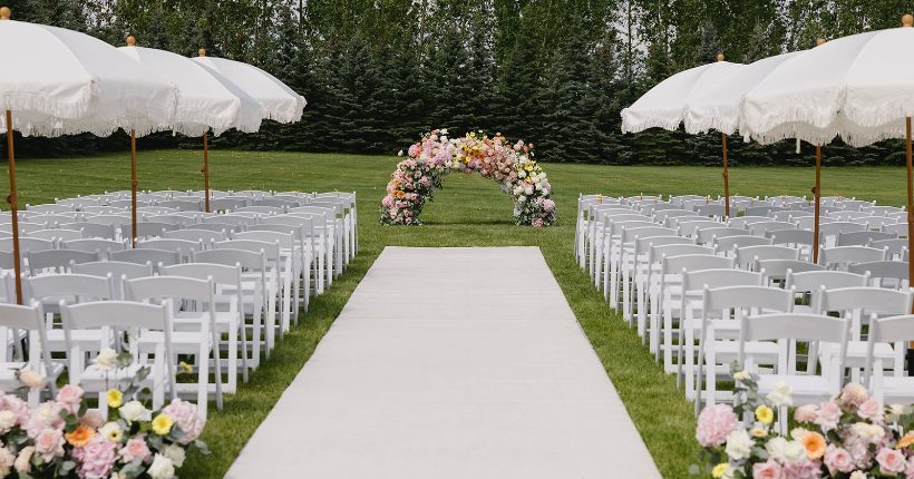 A low floral arch in pink, peach, yellow, at the end of an aisle with neat lines of white chairs and large garden umbrellas.