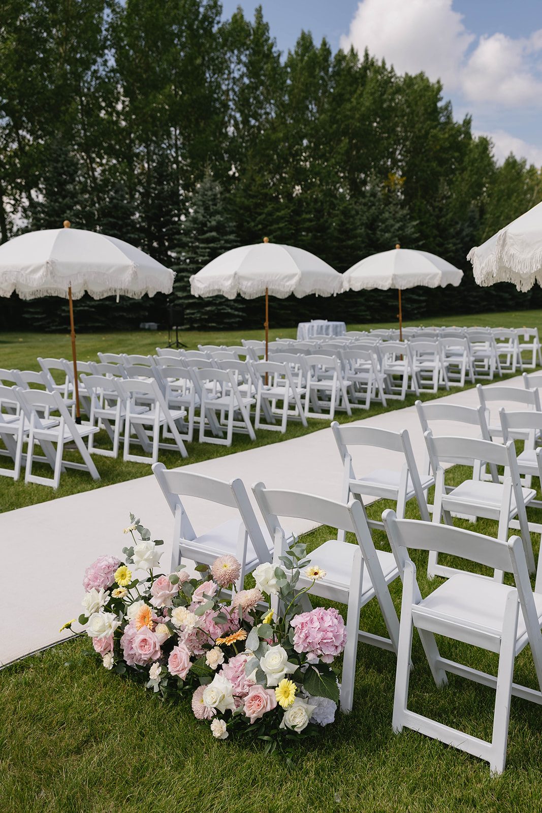 The outdoor ceremony set up for a pastel garden wedding with white chairs in neat lines, large umbrellas lining the aisle, and pastel floral arrangements marking the beginning of the aisle.