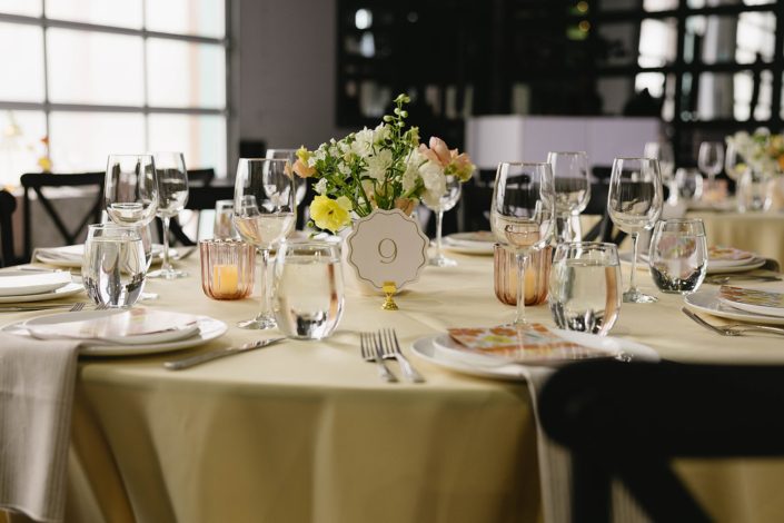 Round guest tables at Tangled Roots indoor reception space with pale yellow linens, a loose, garden inspired centrepiece, and amber glass tea lights.
