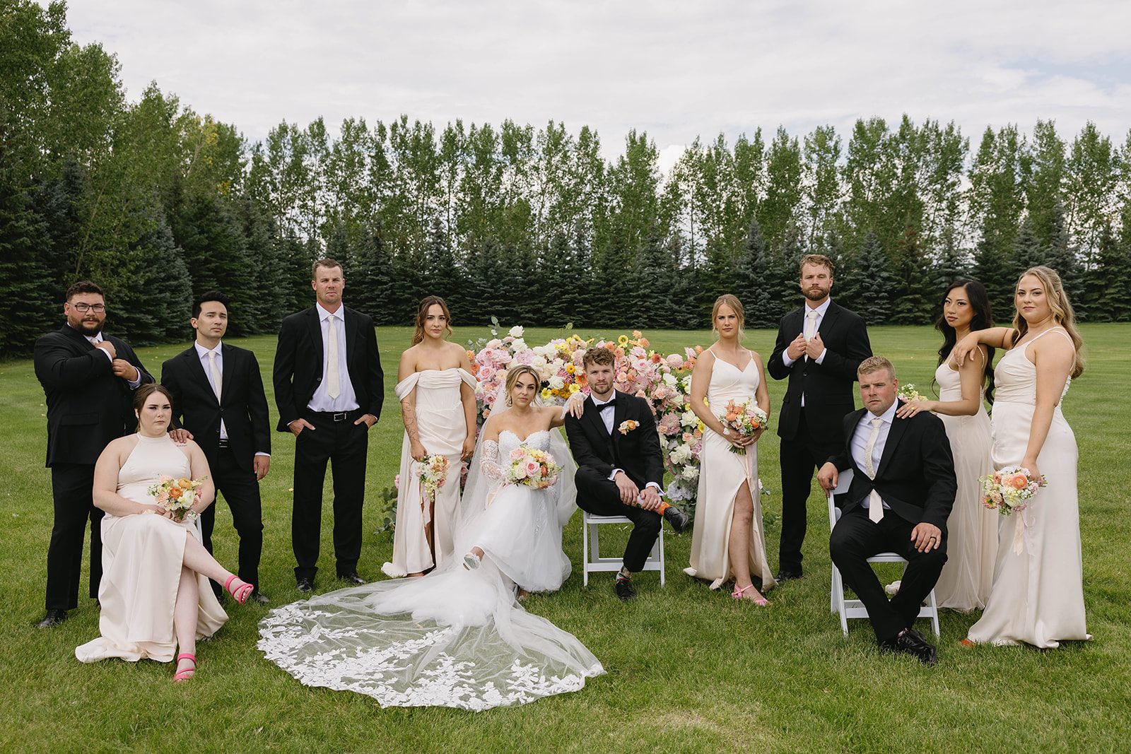 A pastel wedding portrait on the grounds of Tangled Roots in Edmonton featuring a wedding party wearing black suits and neutral toned dresses, the women carrying pastel bouquets.