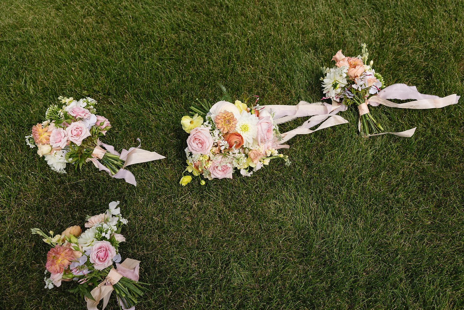 Pastel bouquets in warm tones featuring dahlias, ranunculus, garden roses, stock, lisianthus, sweet peas and orlaya with trailing silk ribbons laying in the grass.