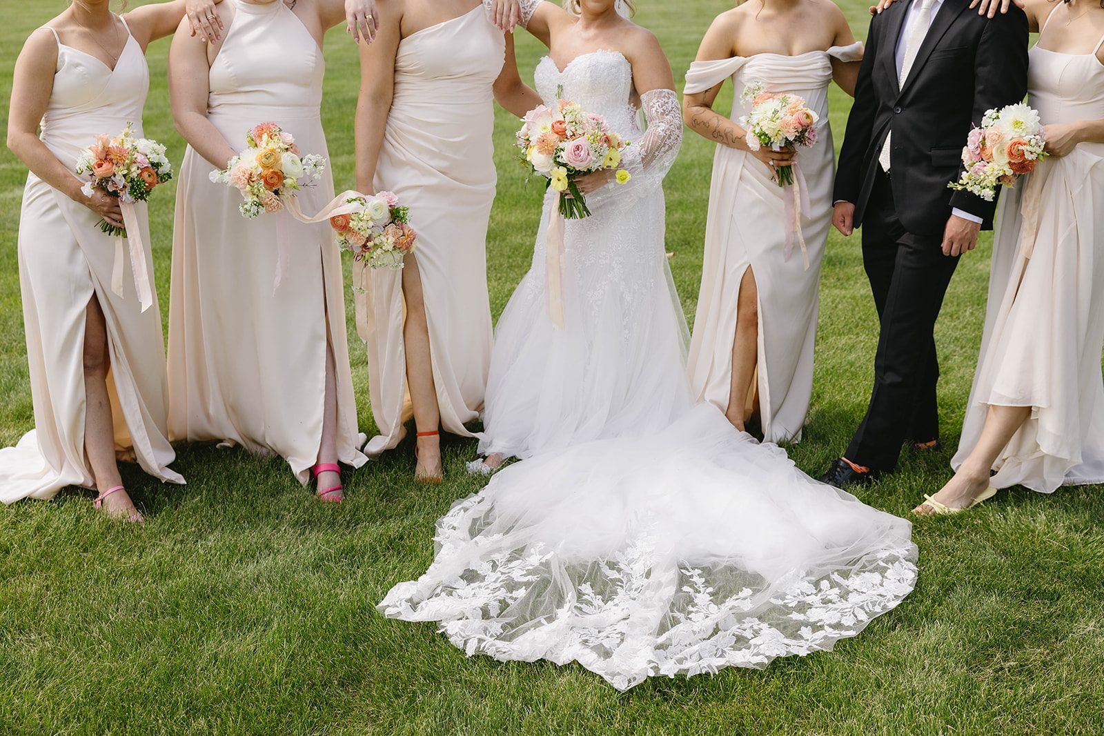 Bride and her bridesmaids in neutral toned dresses holding pastel garden inspired bouquets in blush pink, apricot, peach, and butter yellow tones with trailing silk ribbons.