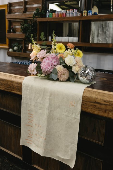 At the bar of Tangled Roots indoor reception space, a pastel arrangement of hydrangea, ceremonies, gerbera daisies, stock, and ranunculus accented by some small disco balls.