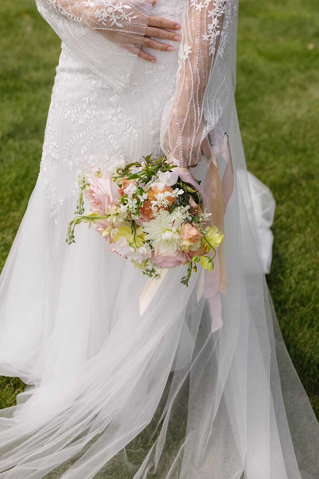 A bride holds a bouquet of garden roses, dahlias, lisianthus, stock, ranunculus, sweet peas and orlaya in blush pink, peach, butter yellow with trailing silk ribbons.