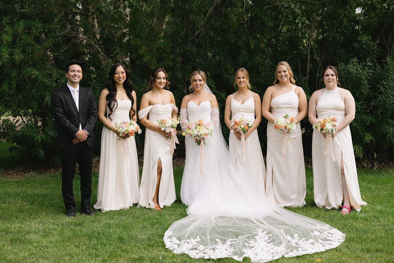 A bride and her bridal party stand at the edge of the trees at Tangled Roots property holding pastel warm toned bouquets of dahlias, garden roses, ranunculus, stock, sweet peas, and orlaya.