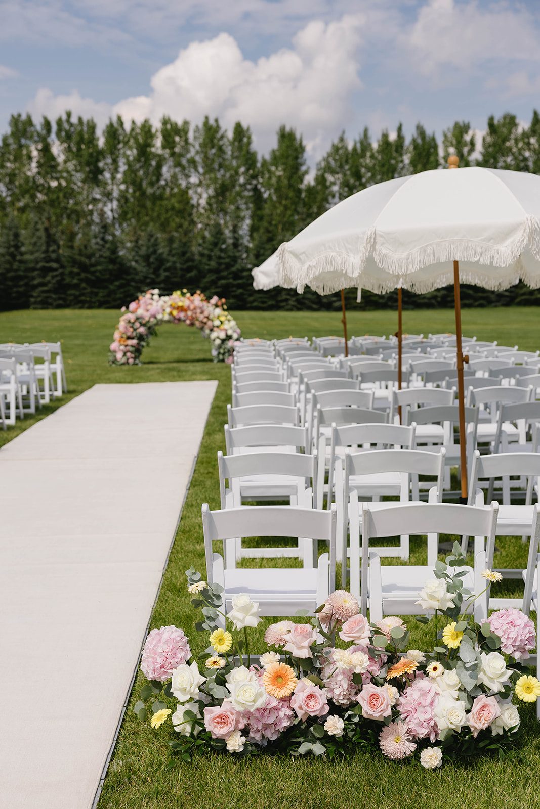 Pretty pastel garden wedding outdoor ceremony at Tangled roots featuring pretty pastel floral arrangements, a low, ombre floral arch, neat rows of white chairs and several large white umbrellas lining the aisle.