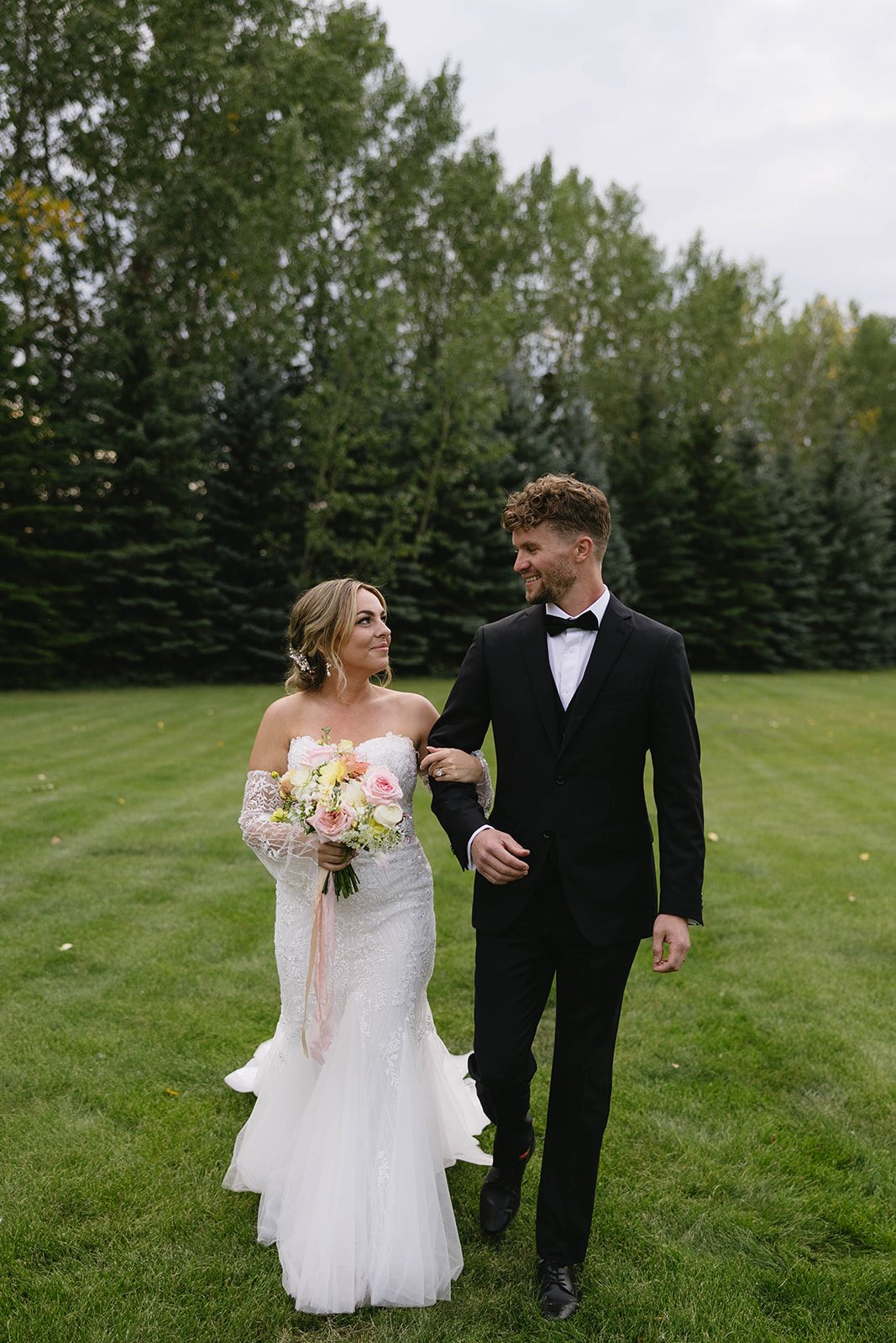 Bride and groom walking arm in arm through the Tangled Roots grounds, looking at each other and smiling. Bride carries a pastel garden inspired bouquet with trailing silk ribbons.