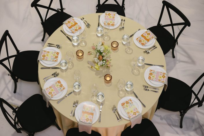 A round guest table viewed from above showing the pale yellow table linens, striped napkins, floral stationery at each place setting, and a pastel garden inspired centrepiece accented by amber glass tea lights.